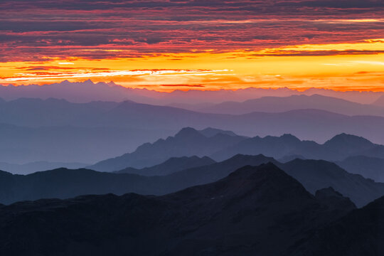 Layered mountain ranges fade into the distance under a vibrant orange and red sunset. Mantova refuge al Vioz, Peio, Trentino. The sky is streaked with colorful clouds.