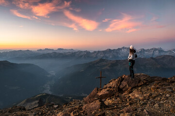 A woman stands on a rocky peak at sunset, overlooking a vast mountain range. A wooden cross stands nearby. Mantova refuge al Vioz, Peio, Trentino. The sky is a blend of orange, pink, and blue hues.