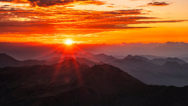 A vibrant orange sunset over layered mountain ranges. The sun bursts through the clouds, casting rays down the peaks. Mantova refuge al Vioz, Peio, Trentino. The view from the refuge is breathtaking.