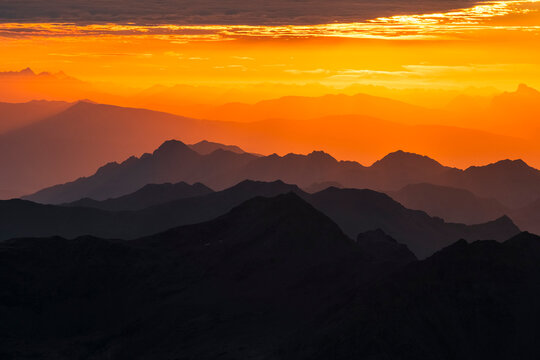 Layers of dark mountain silhouettes fade into an orange sky at sunset. The sky is bright orange with some clouds at the top. Mantova refuge al Vioz, Peio, Trentino 
