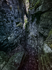 A narrow path winds through a dark, rocky crevice with moss-covered walls. Natural light filters in, highlighting textures of stone and green moss in a mysterious, serene landscape.