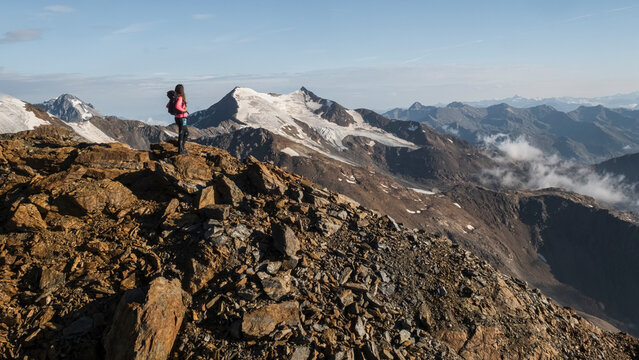 A woman with a backpack stands on a rocky mountain peak, gazing at a vast mountain range with glaciers and clouds. Mantova refuge al Vioz, Peio, Trentino. The hiker is near the refuge.