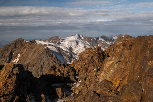 A high-angle shot of rocky, brown mountains with snow-capped peaks in the distance under a cloudy sky. View from the top  of Vioz Mount, Trentino.