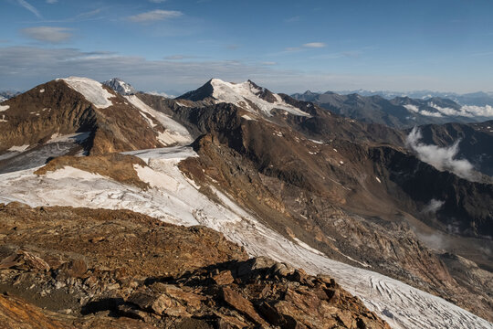 Mountain range with snow-capped peaks under a blue sky with scattered clouds. Brown rocky terrain in the foreground. View from the top  of Vioz Mount, Trentino.