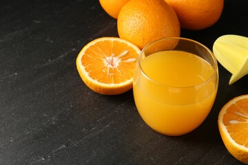 Fresh orange juice in glass, fruits and juicer on black table, closeup. Space for text