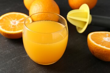 Fresh orange juice in glass, fruits and juicer on black table, closeup