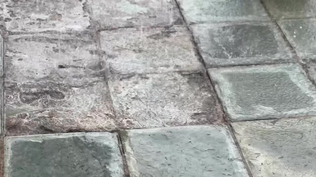 Water pours from a spout onto a tiled concrete floor, creating ripples and puddles and flowing across the textured surface.