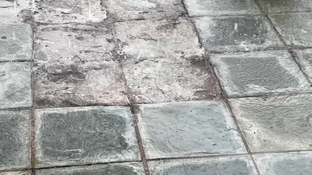 Water pours slow motion from a spout onto a tiled concrete floor, creating ripples and puddles and flowing across the textured surface.