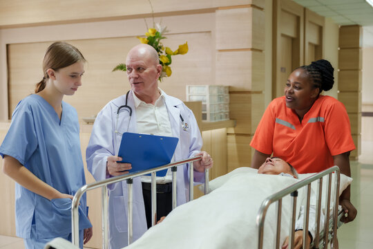 Caucasian senior doctor reviewing treatment file with young female assistant beside hospital stretcher while african female caregiver attending patient during emergency medical discussion in hallway - Powered by Adobe