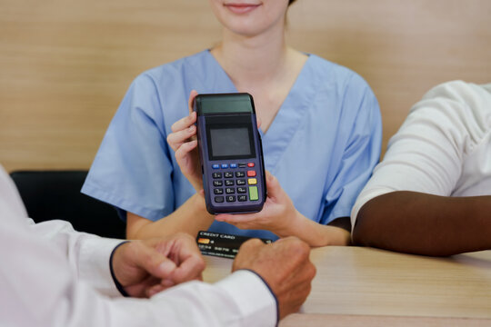 Caucasian female nurse showing payment terminal to senior asian male patient holding credit card for completing transaction at hospital reception desk during medical billing process