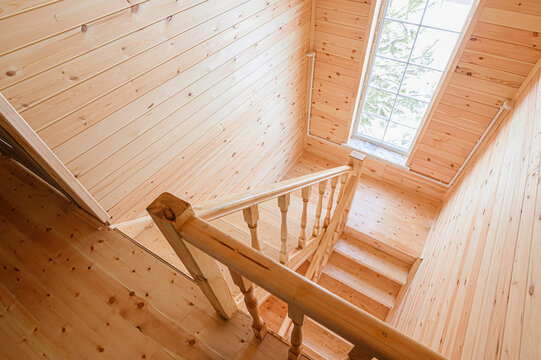 wooden staircase leads down in a room with walls and ceiling clad in light wood paneling. A window lets in natural light