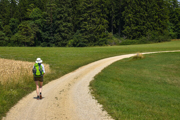 A woman with a bright green backpack walks along a rural path through golden Swiss fields on a sunny summer day