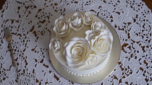 An elegant cake adorned with white roses, placed on a lace doily with a fork in the periphery, hinting at a celebration.