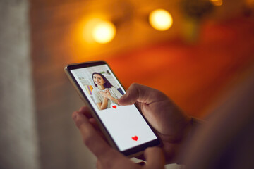 Find love online concept. Man holding smartphone, looking at attractive young woman's profile photo on social media or mobile dating app and pressing red heart like button. Close-up, blur, soft focus