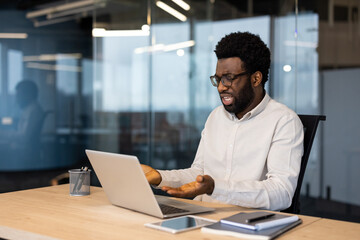 Fototapeta premium A professional man expressing frustration while seated at a desk with a laptop in a contemporary office setting. The image conveys workplace stress and challenges in a high-tech environment.