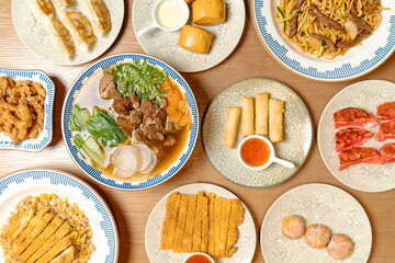 Top view of various Asian dishes including noodles, soup, spring rolls, fried rice, gyoza, and desserts arranged neatly on ceramic plates over a wooden table.