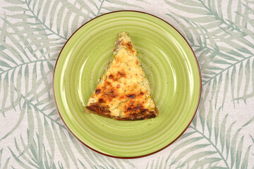 A savory slice of zucchini and leek quiche is centered on a green ceramic plate with a brown rim, set against a leafy patterned tablecloth