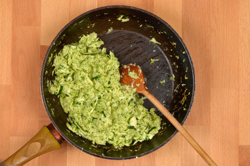 A dark frying pan contains grated zucchini being stirred by a wooden spoon, resting on a light-colored wooden background