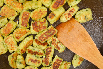 A close-up shot shows a wooden spatula flipping fried zucchini gnocchi with a golden-brown crust in a dark pan