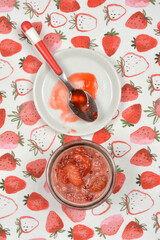 A top-down view of a jar of homemade strawberry jam and a small plate with a tasting spoon on a fabric with a strawberry pattern
