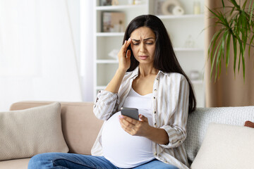 Pregnant woman looking worried while checking smartphone at home. Reflects anxiety or stress, possibly related to pregnancy health or digital information overload