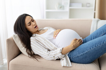 Pregnant woman lying on sofa holding her belly, looking distressed. Captures a contraction, labor pain or realistic symptom of late stage pregnancy, closeup