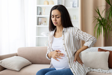 Obraz premium Pregnant woman experiencing back pain and fatigue while sitting on sofa, holding her belly and side. Realistic view of physical discomfort during pregnancy, closeup