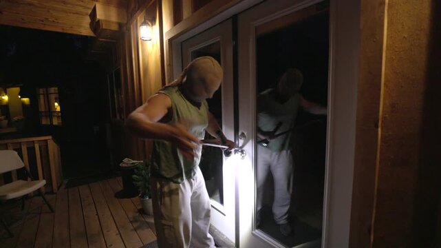 masked man holding a crowbar and looking into a home through window blinds at night. The unsettling atmosphere evokes fear and suspense, ideal for illustrating concepts of crime, danger, home invasion