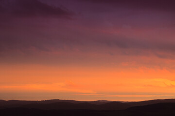 Obraz premium A panoramic view of rolling hills in silhouette against a dramatic sunset sky with a gradient of vibrant orange and deep purple colors.