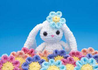 Close up of hand crochet Easter Bunny with crochet flower over ear and in front of bunny, on a blue background.
