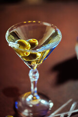 A martini glass with olives, resting on a wooden surface with a blurred, warm-lit background.