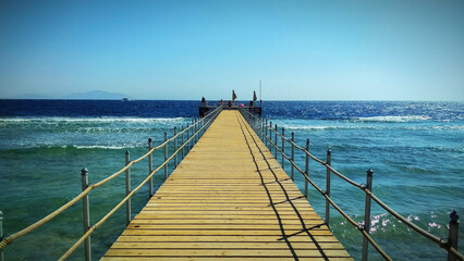 Fototapeta premium Long wooden pier leading into the clear turquoise sea under a bright blue sky. Idyllic vacation setting with a scenic view of the water and distant horizon.