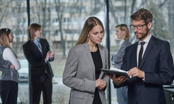 Two people in suits are standing in a room, one of them holding a clipboard
