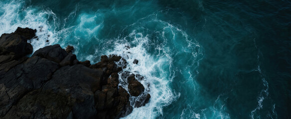 Rocky cliffside with deep blue ocean waves crashing against brown rocks. White foamy water, trees on cliff provide contrast to wild ocean nature. Blue, white colors with warm brown rock tones.