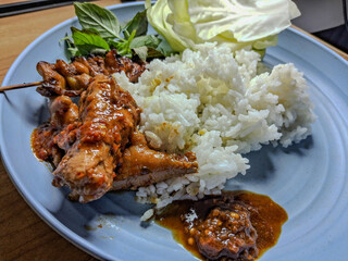 Traditional Indonesian meal with rice, grilled chicken satay, sambal, raw cabbage, and fresh basil leaves served on a blue plate