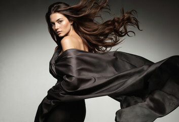 Naklejka premium Woman with long flowing hair stands in dynamic pose against light gray backdrop. Black dress and caught hair mid-motion add energy and movement. Gaze directed to left draws attention.