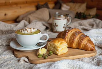 Breakfast scene on wooden table in log cabin setting. Warm coffee mug with whipped cream and croissant on wooden board. Green plant and single flower vase add rustic charm to cozy atmosphere.