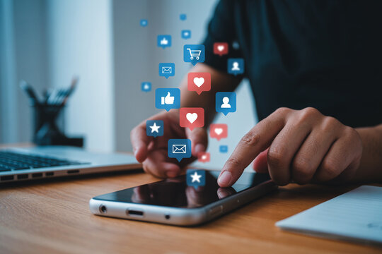 Hands using a smartphone surrounded by social media icons for likes comments and shares on a wooden desk top with a laptop nearby