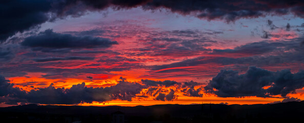 A breathtaking panoramic view of a fiery red and orange sunset with dark, textured clouds stretched across the sky over a mountain range.