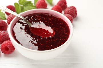 Sweet raspberry jam in bowl, berries and green leaves on white wooden table, closeup. Space for text