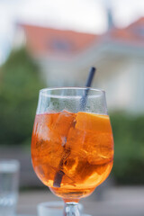 A vibrant orange Aperol Spritz cocktail, garnished with a slice of orange, sits in a glass with ice on a sunny terrace.