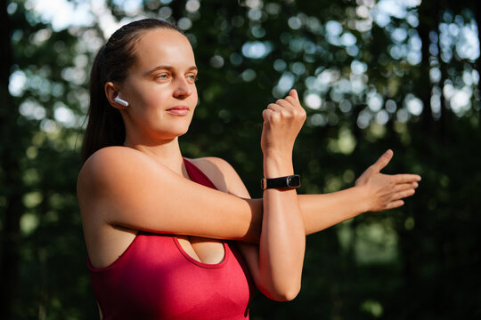 A young woman in a red tank top with a ponytail, earbuds, and a fitness tracker stretches her arm across her body in a sunlit, wooded park.