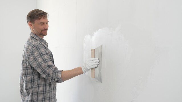 Man construction worker wearing protective gloves and plaid shirt, spreading plaster smoothly across wall surface using professional drywall taping knife during renovation project