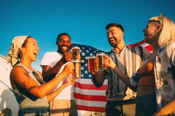 Cheerful multi-ethnic friends toasting with beer mugs celebrating with usa flag on rooftop party at sunset
