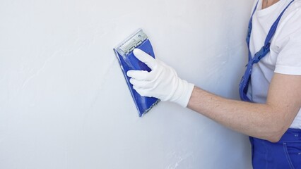 Unknown male construction worker wearing protective white gloves and blue construction coveralls, is sanding white wall with blue hand block, preparing smooth surface during interior home renovation © volha_r