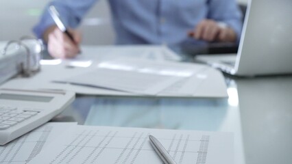 Calculator, pen and documents are lying on accountant's desk. Businessman wearing blue shirt is working with documents on the background. Business people, audit concept