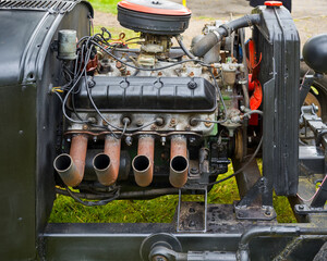 Close-up of a vintage car engine with visible exhaust pipes, wiring, and carburetor. The engine is partially disassembled, showcasing mechanical components and a patina of age and use.