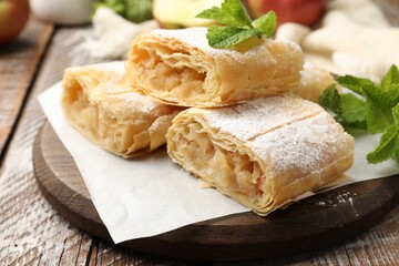 Pieces of tasty apple strudel with powdered sugar, mint and fruits on wooden table, closeup