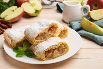 Pieces of tasty apple strudel with powdered sugar and mint on light wooden table, closeup