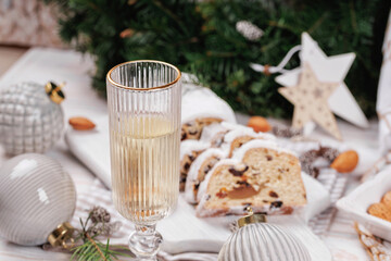 A tall champagne glass filled with sparkling wine stands as the centerpiece on a Christmas table surrounded by stollen bread, pinecones, ornaments, and festive greenery.